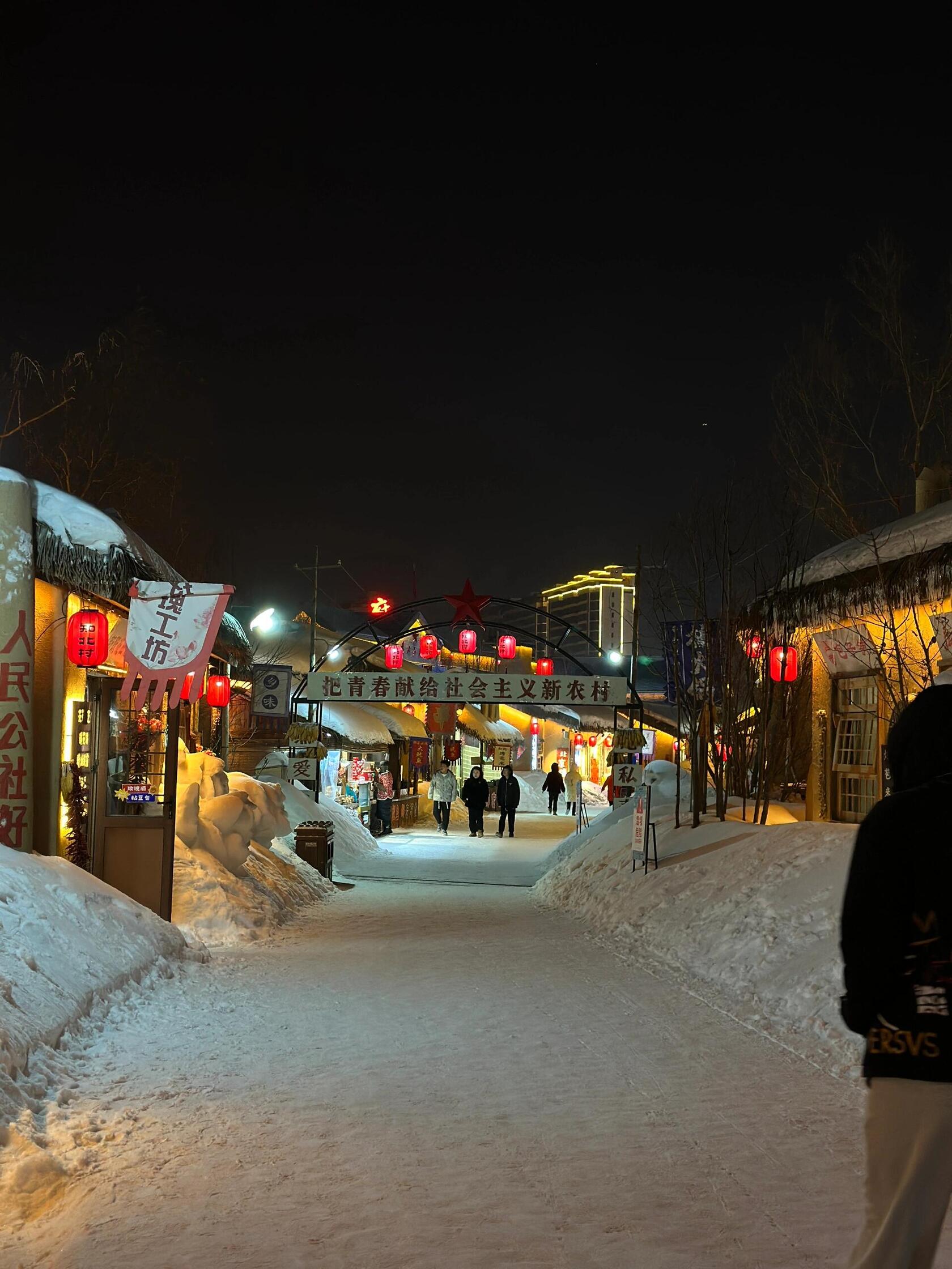 A snow-covered street in a traditional Chinese village at night, illuminated by red lanterns.