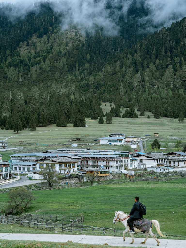 A person riding a white horse through a charming Tibetan village, showcasing the local life on a Tibet self-drive adventure.