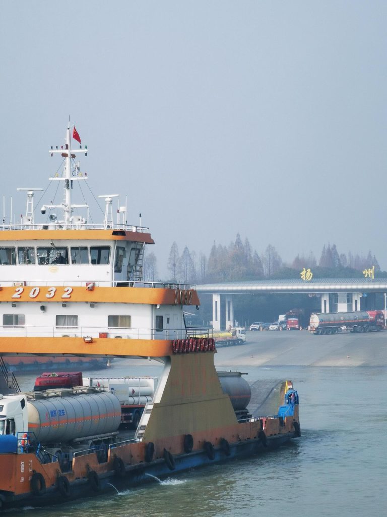 A ferry boat with trucks crossing a river near a bridge with 'Yangzhou' written on it, showcasing transport in Eastern China Exploration.