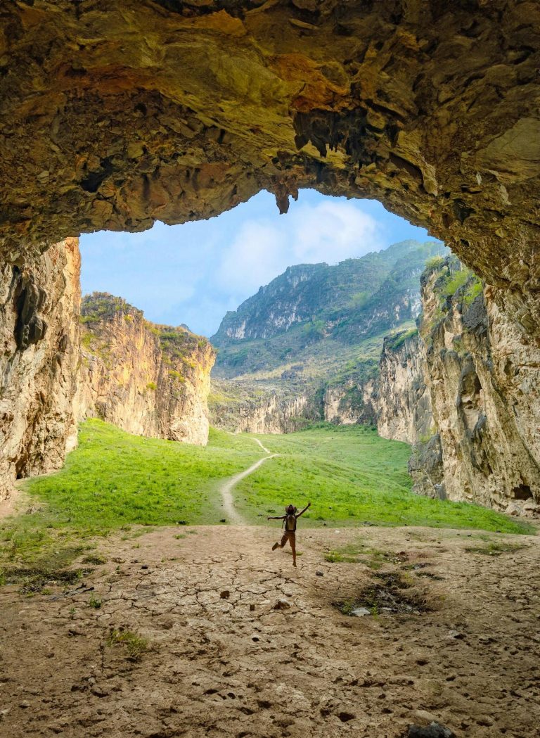 A person jumping in front of a cave entrance overlooking a vast mountain valley, a thrilling Guangdong Nature Adventure moment.