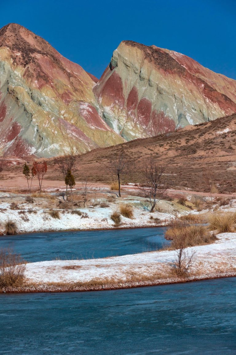 A vibrant, colorful mountain range with a deep blue river, set against a clear blue sky, showcasing Gansu's stunning geology.